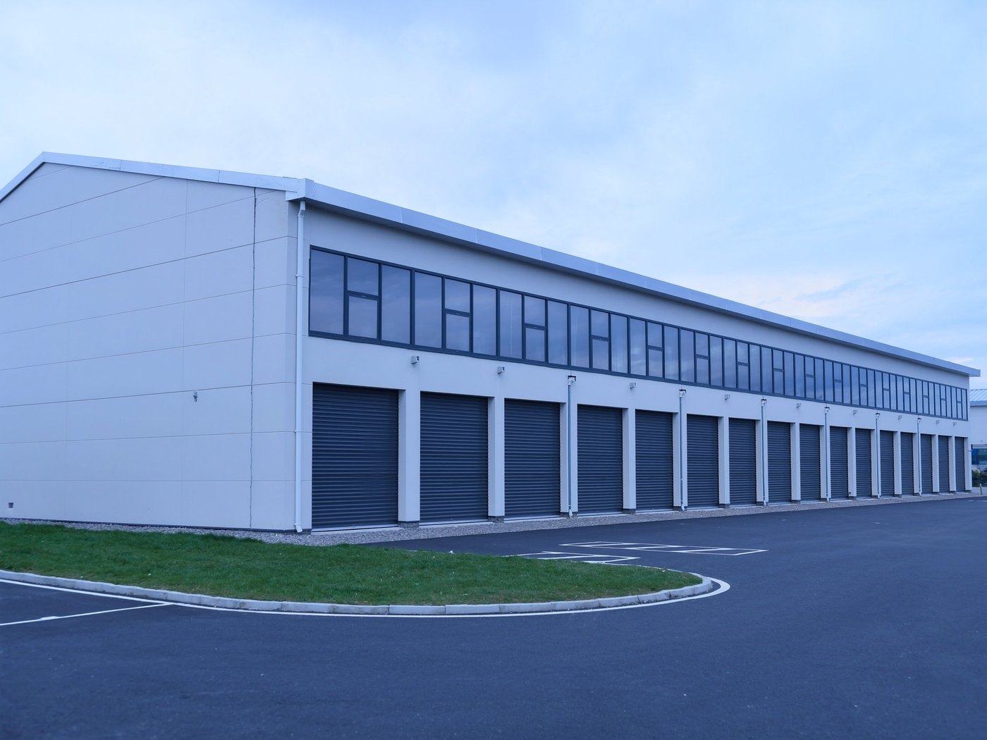 Wide view of a completed self-storage facility: a long pale metal-clad facade, a row of closed steel roller doors, a glazed clerestory band above, and asphalt hardstanding in the foreground